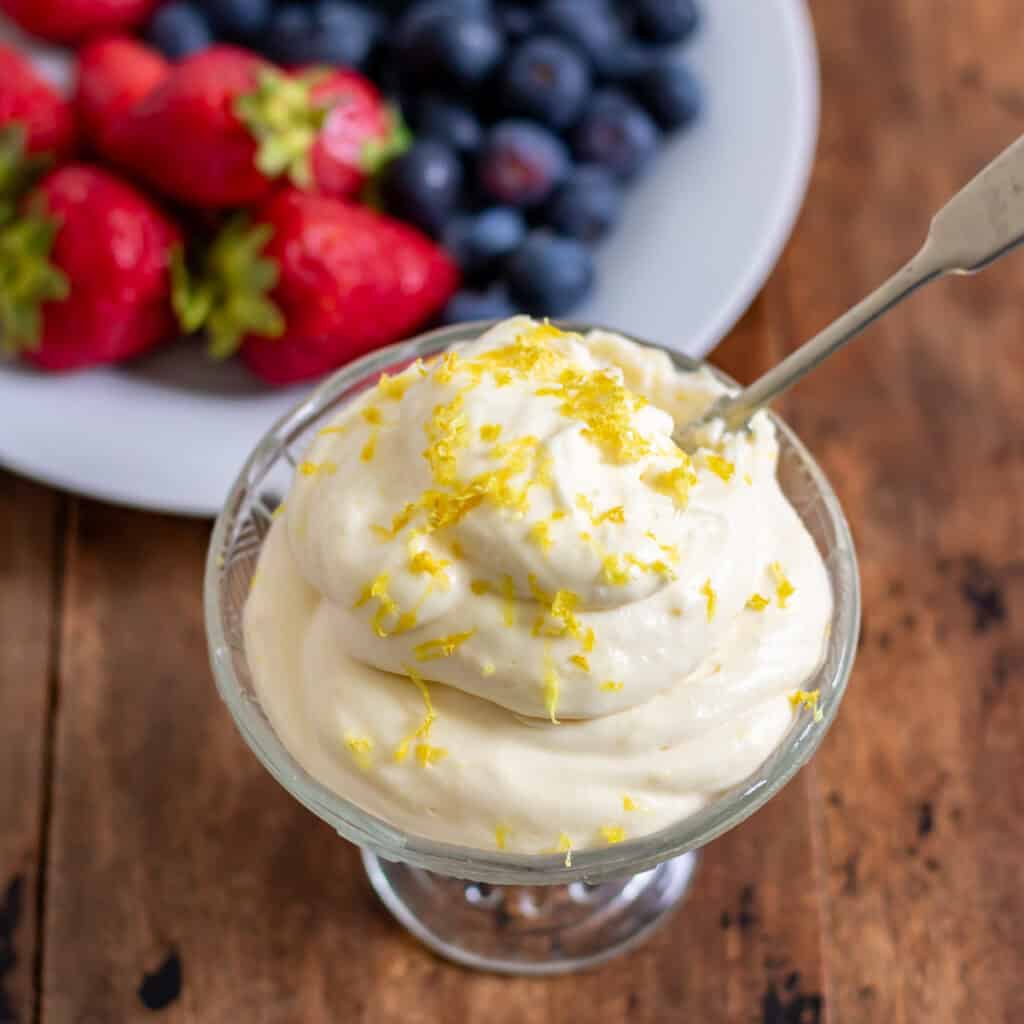 A glass dish piled with lemon whipped cream, topped with extra lemon zest, in front of a dish of berries.