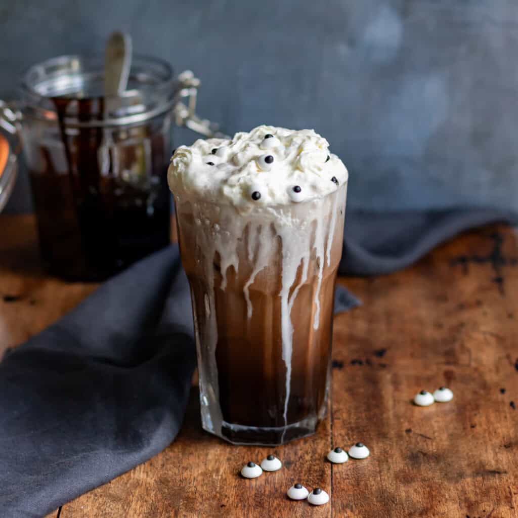 A glass of Halloween chocolate cream soda, topped with whipped cream and candy eyes, with cream drizzling down the outside of the glass.